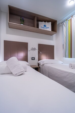 Ein Hotelzimmer mit zwei Betten, einem Holzregal darüber mit einer Topfpflanze und einer Club del Sole Tasche, einer Nachttischlampe und einem Fenster mit gestreiftem Vorhang.
