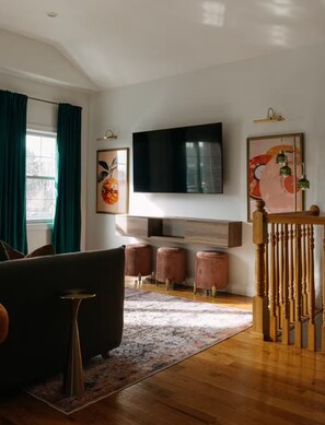 A modern living room with a large flat-screen TV mounted on the wall, a dark sofa, a patterned rug, and a wooden staircase.