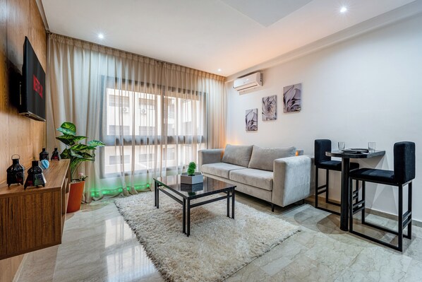 A modern living room with a sofa, coffee table, and two bar stools. There is a flat-screen TV mounted on the wall, a potted plant, and artwork on the wall. - Le Grand Chic - Downtown swimming pool (Marrakesh)