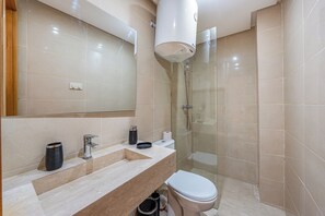 A modern bathroom with a beige tiled wall, a large mirror, a sink with a marble countertop, a toilet, and a glass-enclosed shower area. - Le Grand Chic - Downtown swimming pool (Marrakesh)