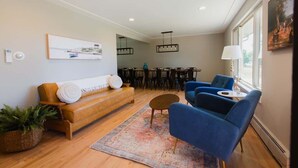 A modern living room with a sofa, blue armchairs, a dining area, and a large window.