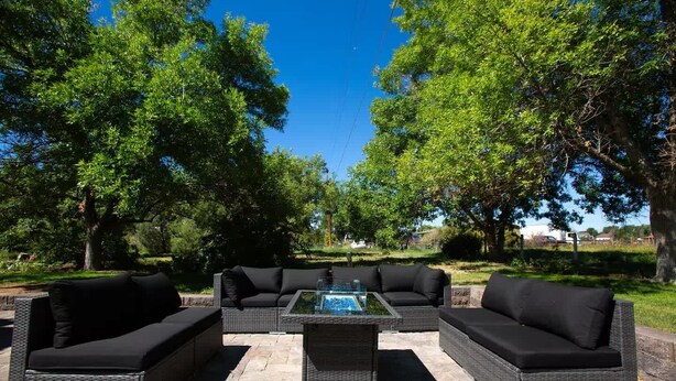 An outdoor seating area with wicker furniture and a glass-top table, surrounded by trees and a clear sky.