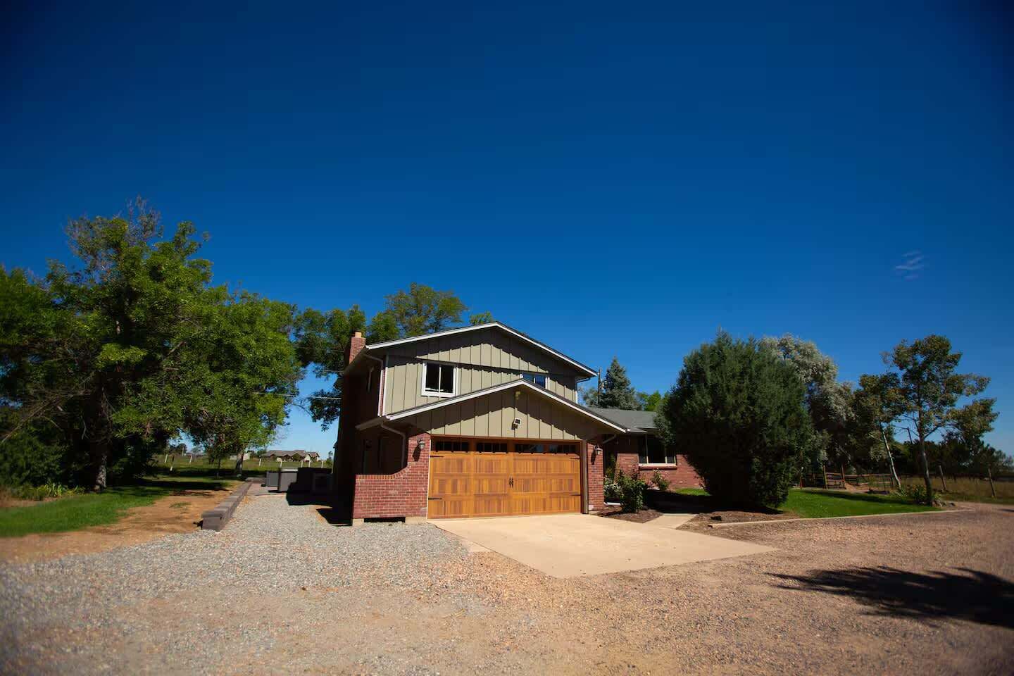 A two-story house with a wooden garage door, surrounded by trees and a clear blue sky.