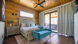 Un dormitorio con techo de madera, una cama con cabecera, un banco, una mesita de noche, una lámpara de pie y una ventana con cortinas.