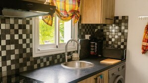 Une cuisine avec un plan de travail noir, des armoires en bois, un four encastré, un lave-linge et une fenêtre agrémentée d’un rideau à carreaux.