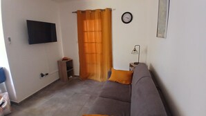 Un salon avec un canapé gris, des coussins orange, un téléviseur à écran plat, une horloge et une lampe sur une table d’appoint.