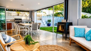 A modern kitchen with a grill, dining area with chairs, a sofa with cushions, and a large window overlooking a garden.