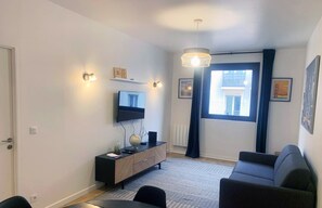 A modern living room with a flat-screen TV, a dark sofa, a dining table, and a patterned rug. - Urban Flat 167 - 1 Bedroom Flat in Paris (París)