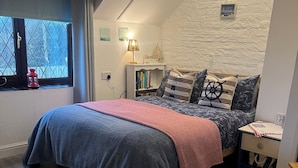 A cozy bedroom with a sloped ceiling, a large bed with a blue and gray duvet, a red throw blanket, and nautical-themed pillows. There is a bedside table with a lamp, a small shelf with books, and a window with a view of trees. - Traditional 1850s fisherman’s cottage, perfect coastal retreat, sea views. (Scratby)