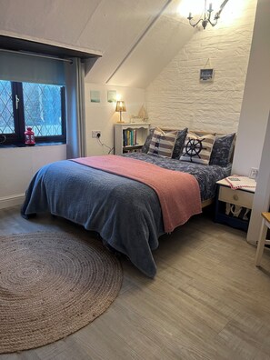 A cozy bedroom with a sloped ceiling, a large bed with a blue and gray duvet, a red throw blanket, and nautical-themed pillows. There is a bedside table with a lamp, a small shelf with books, and a window with a view of trees. - Traditional 1850s fisherman’s cottage, perfect coastal retreat, sea views. (Scratby)