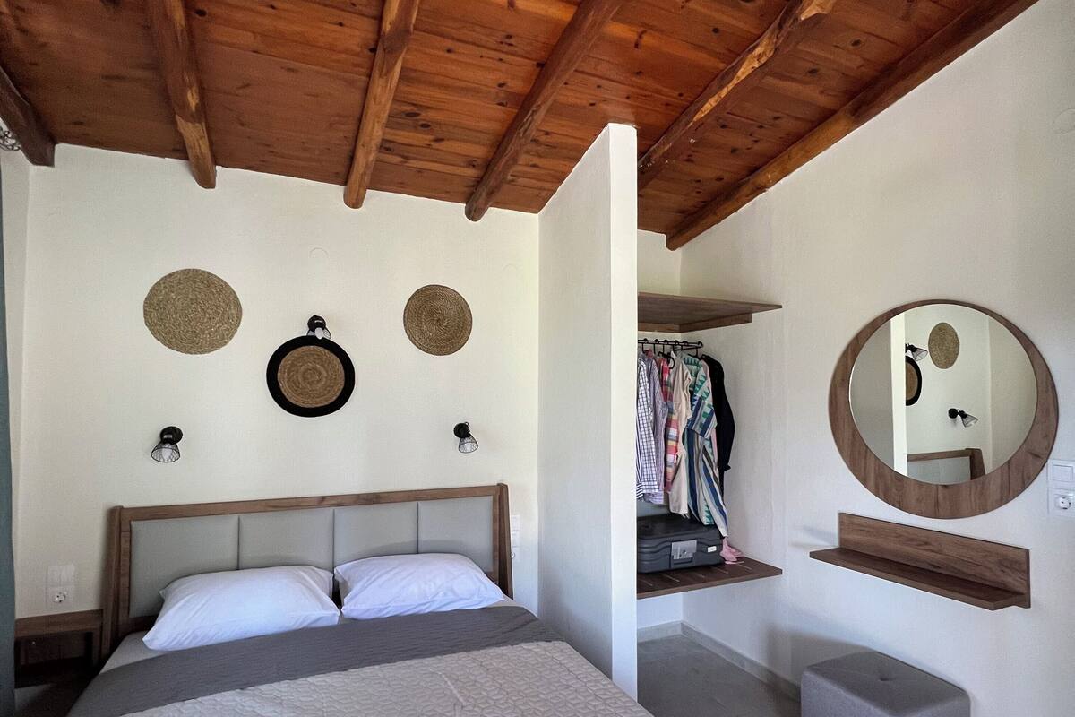 A bedroom with a wooden ceiling, a bed with a grey quilt, a round mirror, and a clothing rack.