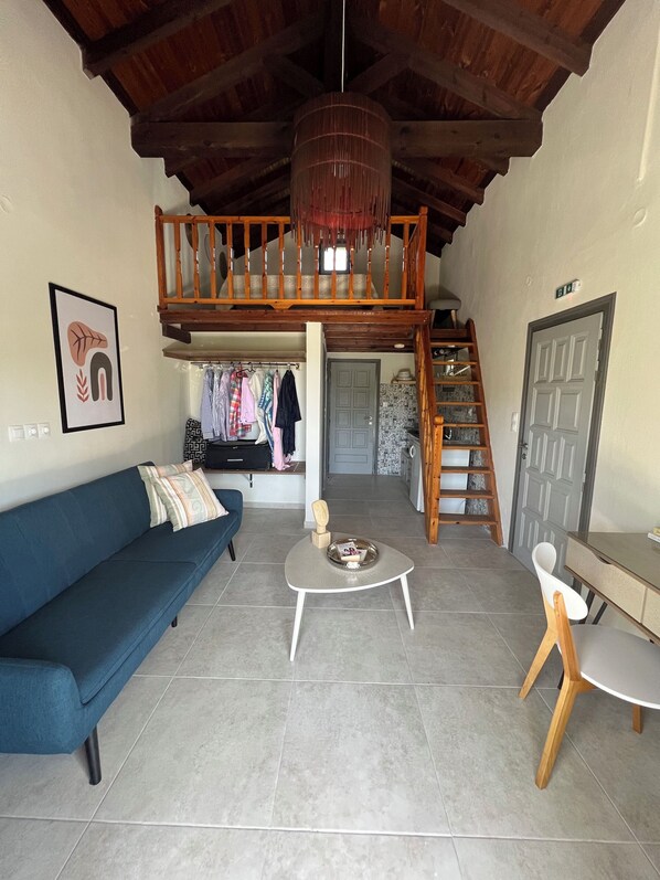 A living room with a blue sofa, a small round table, a white chair, a wooden staircase, and a loft area with clothes hanging. - Ninemo Residences Skiathos (Skiathos)