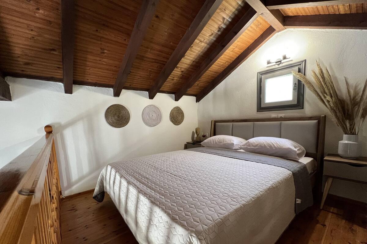 A bedroom with a wooden ceiling, a bed with a quilted bedspread, a nightstand with a vase of dried plants, and a wall-mounted mirror.
