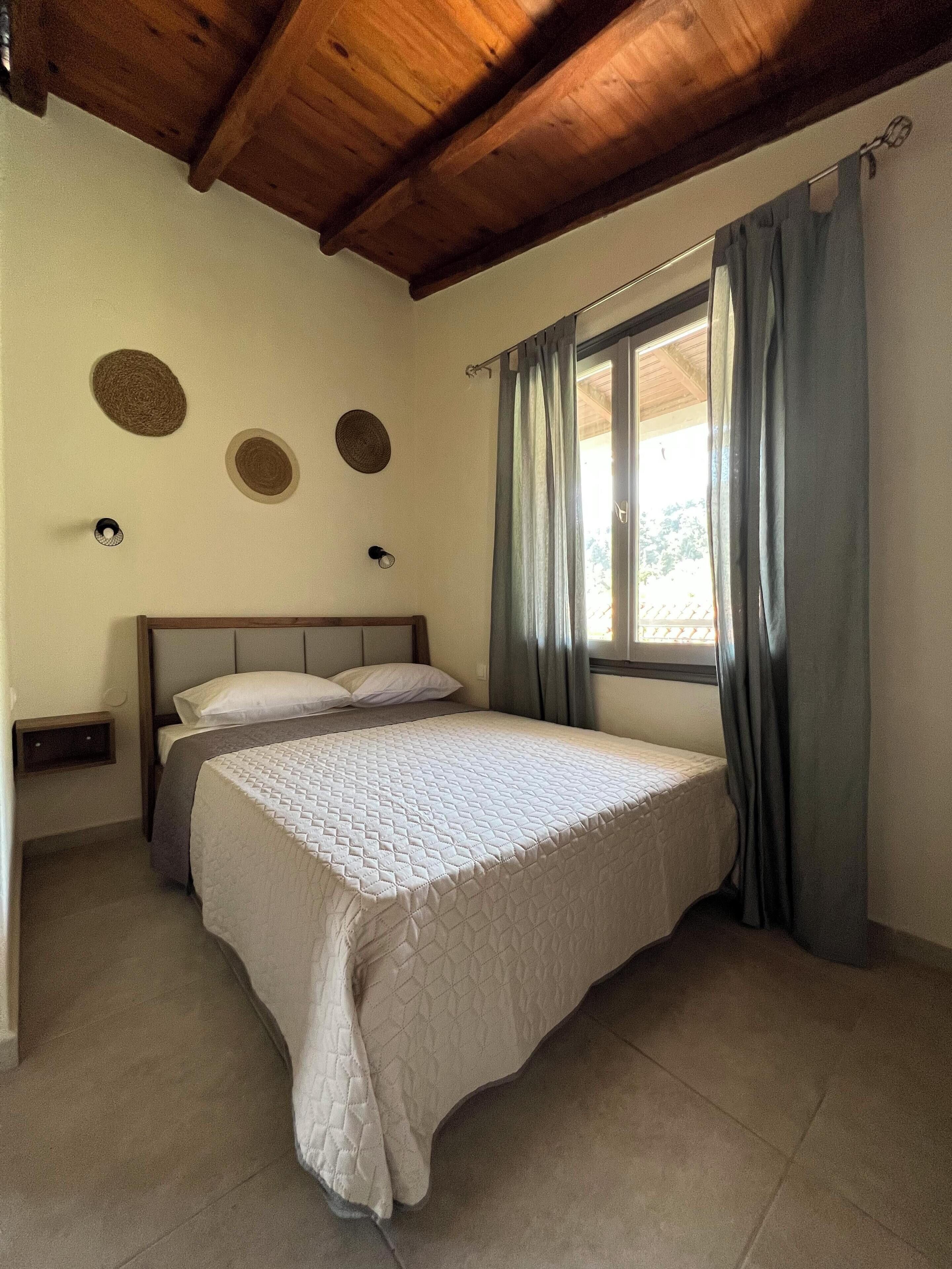 Quarto com teto de madeira, cama com colcha acolchoada, janela com cortinas e detalhes decorativos nas paredes.