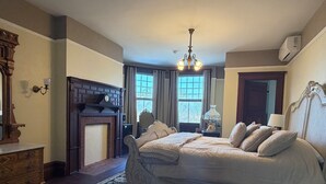 A bedroom with a large bed, a fireplace, a chandelier, and a view of the outdoors through the window.