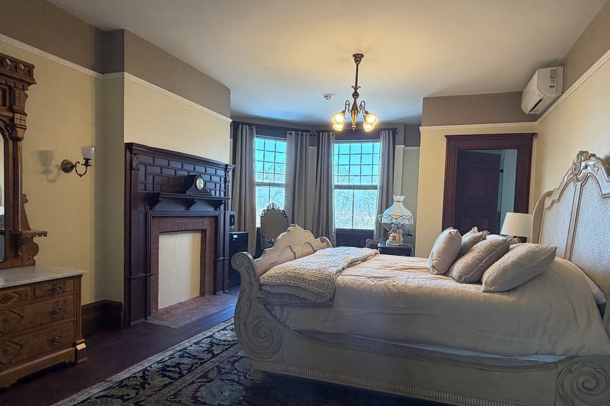 A bedroom with a large bed, a fireplace, a chandelier, and a view of the outdoors through the window.