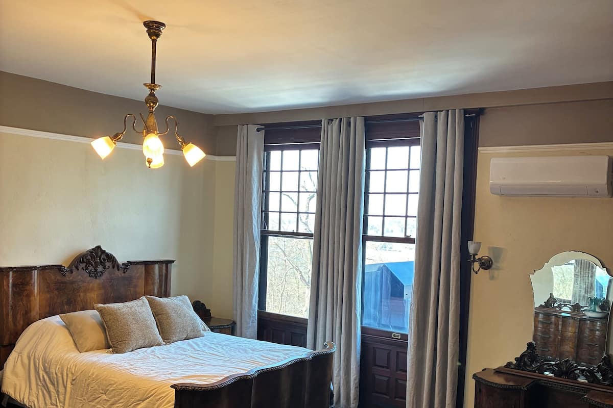 A bedroom with a large bed, a chandelier, a wooden dresser, and a window with curtains.