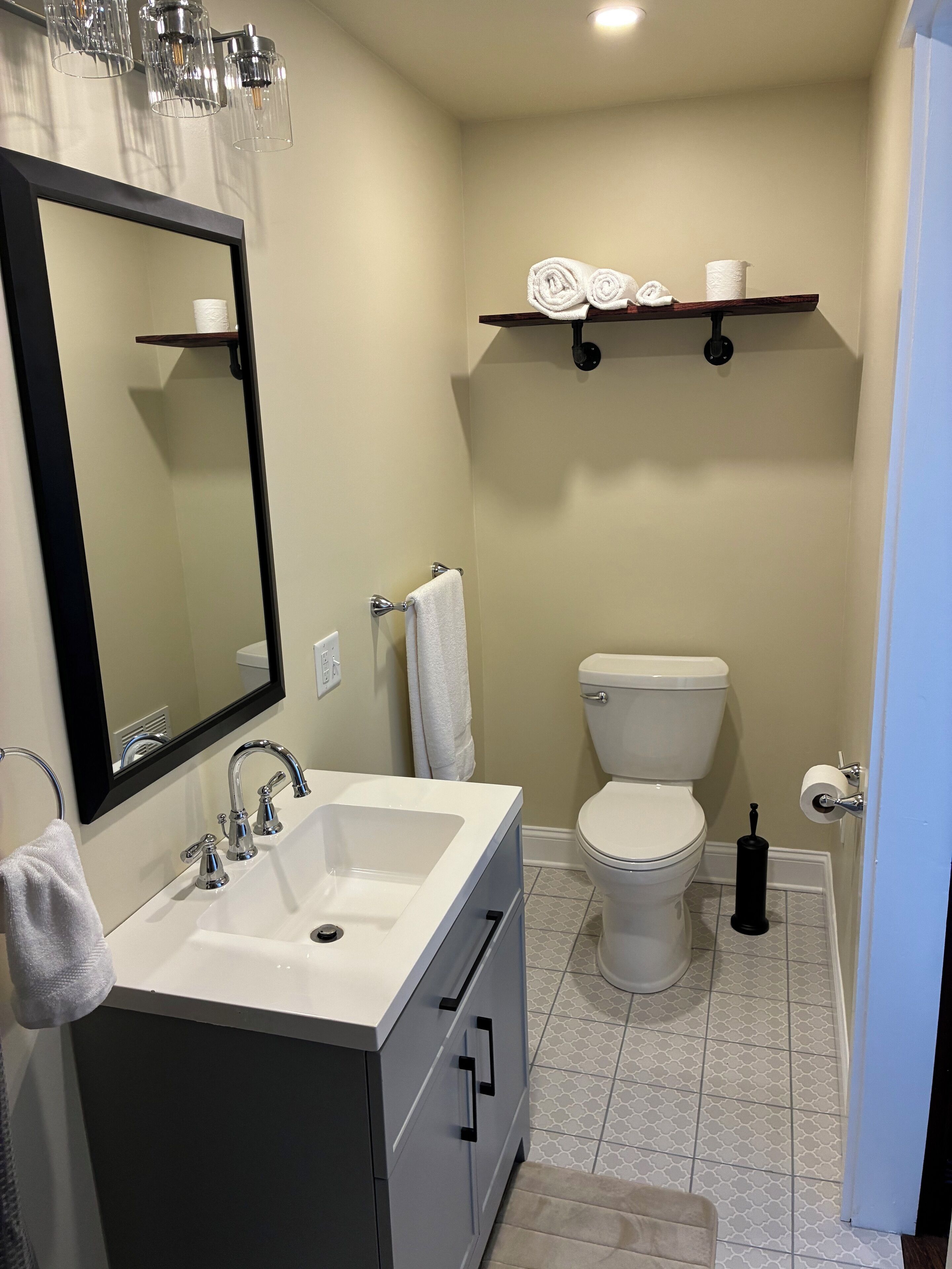 A modern bathroom with a white sink, mirror, and toilet. There are towels and a toilet paper holder on a shelf above the toilet.