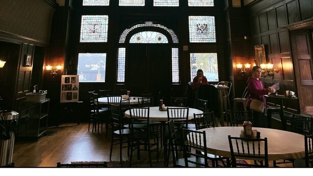 A grand hall with a large chandelier, multiple stained glass windows, and a central arched doorway. Tables and chairs are arranged for dining.