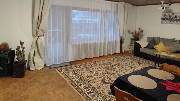 Una sala de estar acogedora con techo de madera, un ventanal con cortinas traslúcidas, un tapete estampado, un sofá con cojines y una mesita auxiliar con adornos.