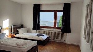 Una habitación de hotel con dos camas, una ventana con cortinas negras, un radiador y suelo de madera.