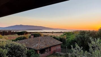 Una casa rustica con tetto in tegole, circondata dal verde e affacciata su uno specchio d'acqua con montagne sullo sfondo.