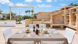 Un área de comedor al aire libre con una mesa dispuesta para cuatro personas, rodeada de estructuras de piedra y sillas blancas.