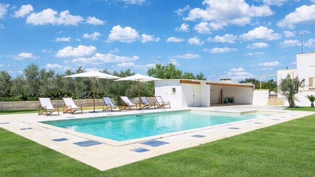 Una piscina con sillones de descanso y sombrillas, rodeada de un césped cuidado y un edificio moderno.