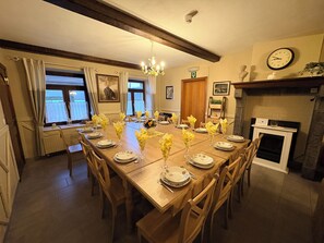 A dining room with a long wooden table set for a meal, flanked by windows with curtains, a chandelier, and a fireplace.