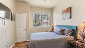 Un dormitorio con cama, mesita de noche, lámpara, televisión y ventana con vista a árboles.