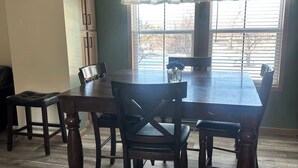 Een eetkamer met een grote houten tafel en stoelen, een raam met luxaflex en een kast met een 'GATHER' bord.