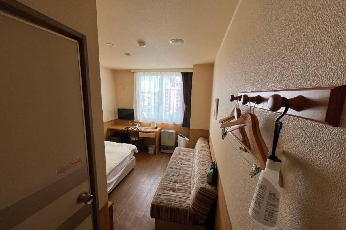 A compact hotel room with a bed, a small desk, a chair, a sofa, and a wall-mounted hook system.