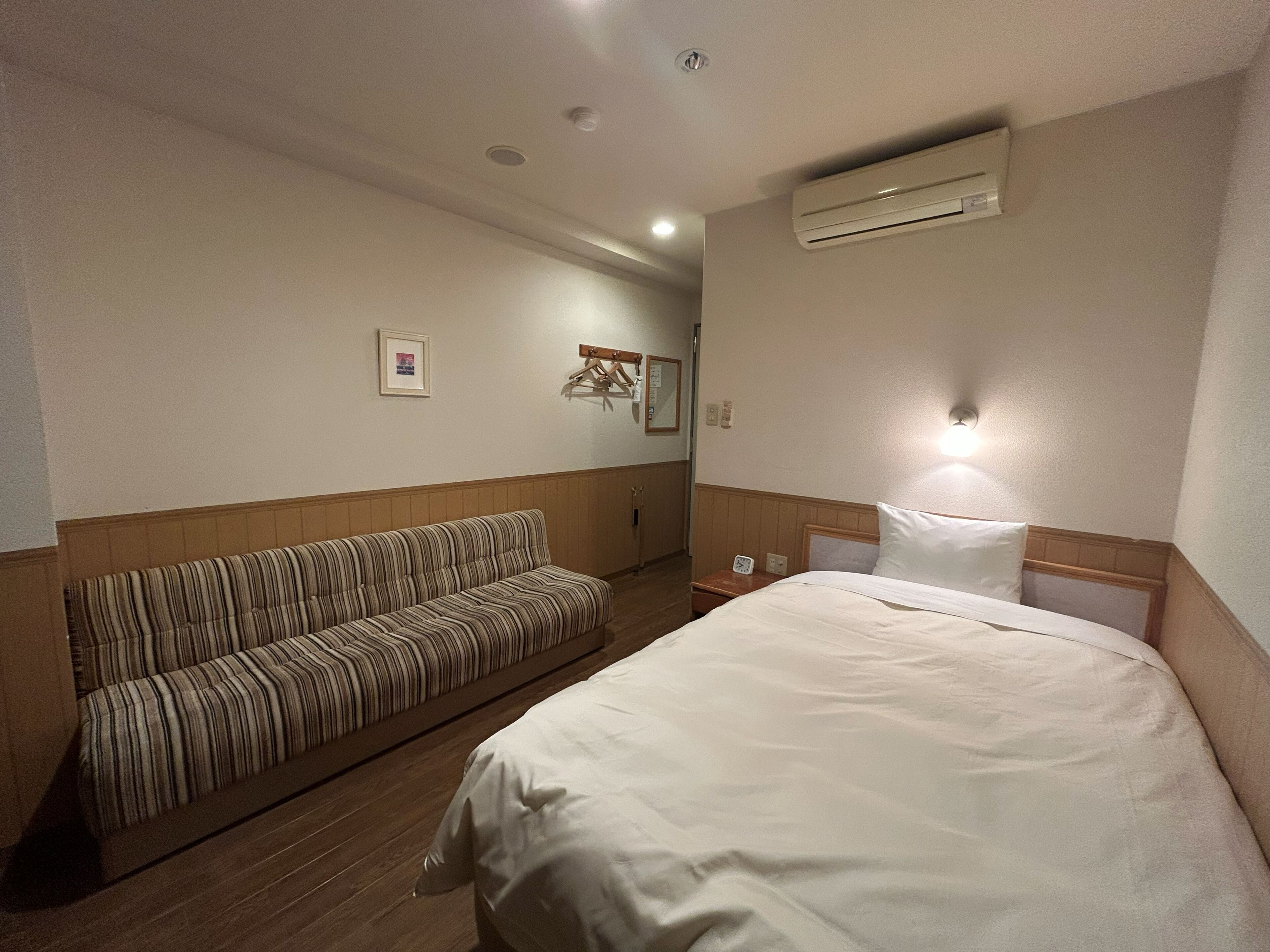 A hotel room with a bed, a striped sofa, an air conditioning unit, a wall-mounted light, and a framed picture.