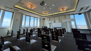 A conference room with rows of chairs, a projector screen, and large windows offering a city view. - Lasting Hotel (Kushiro)