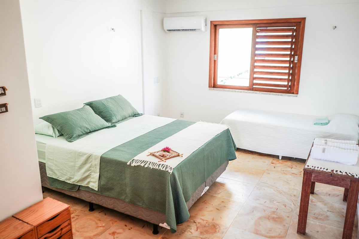 A bedroom with a bed, a bedside table, a window with blinds, and an air conditioning unit.