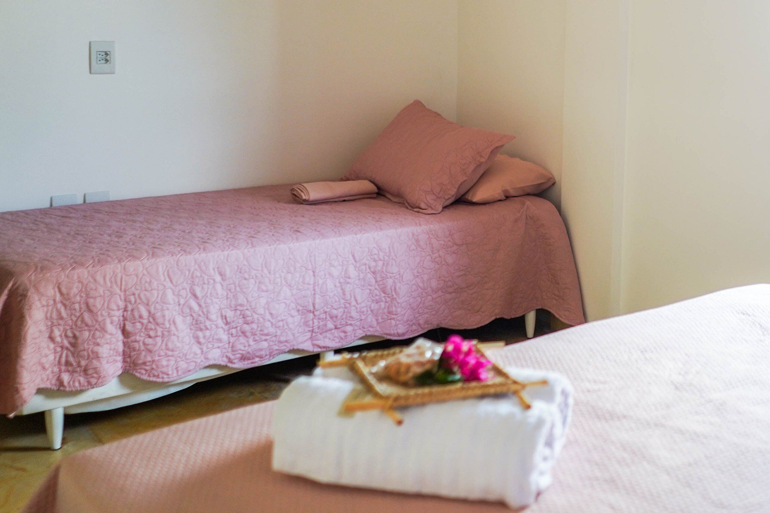 Una cama individual con una colcha y almohadas rosas, una mesita de noche blanca con una toalla blanca doblada y un pequeño objeto decorativo, y una pared lisa con un enchufe.