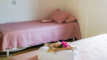 Una cama individual con una colcha y almohadas rosas, una mesita de noche blanca con una toalla blanca doblada y un pequeño objeto decorativo, y una pared lisa con un enchufe.