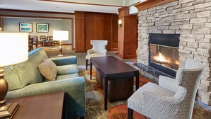 Eine Hotelhalle mit Kamin aus Stein, grünen Sofas, gemusterten Sessel und einem Essbereich mit Tisch und Stühlen.