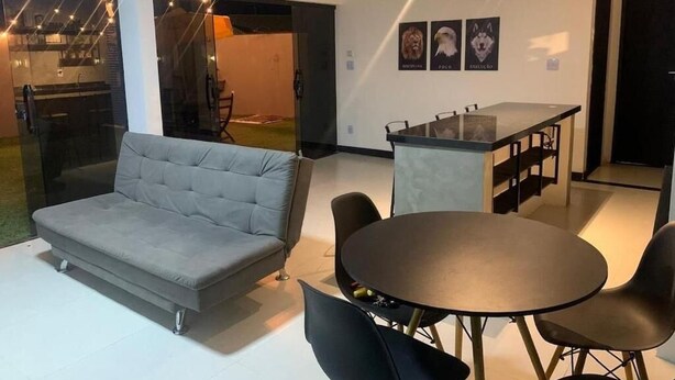 Una sala moderna con un sofá gris, una mesa redonda y sillas negras. Se observa un área de cocina con una barra divisoria a través de una partición de vidrio.