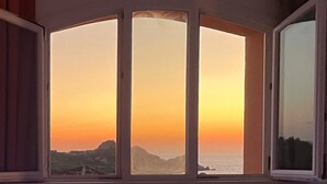 A room with a large window featuring a view of a sunset over a coastal landscape. - Sunset Costarossa 1223 - Breathtaking sea view terrace Sardinia (Trinità D'Agultu e Vignola)