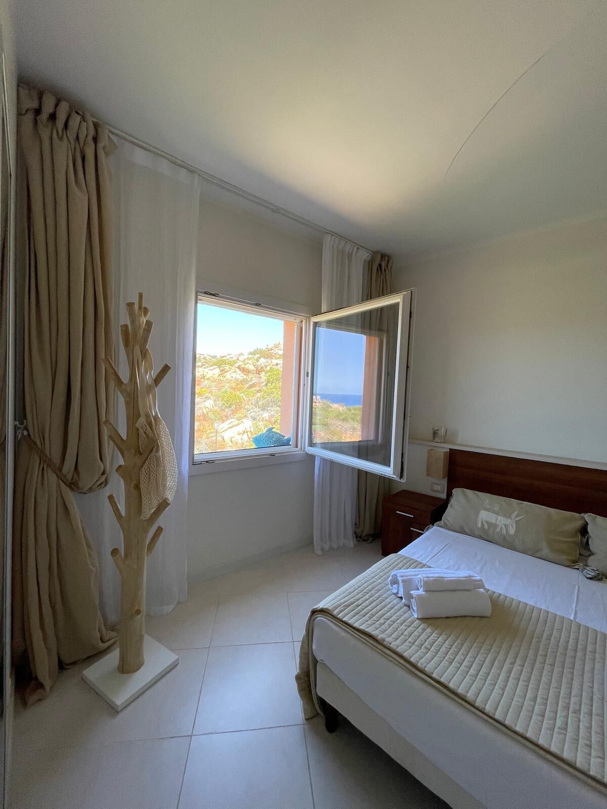 Quarto de hotel com cama, mesa de cabeceira, janela com cortinas e uma escultura de madeira.