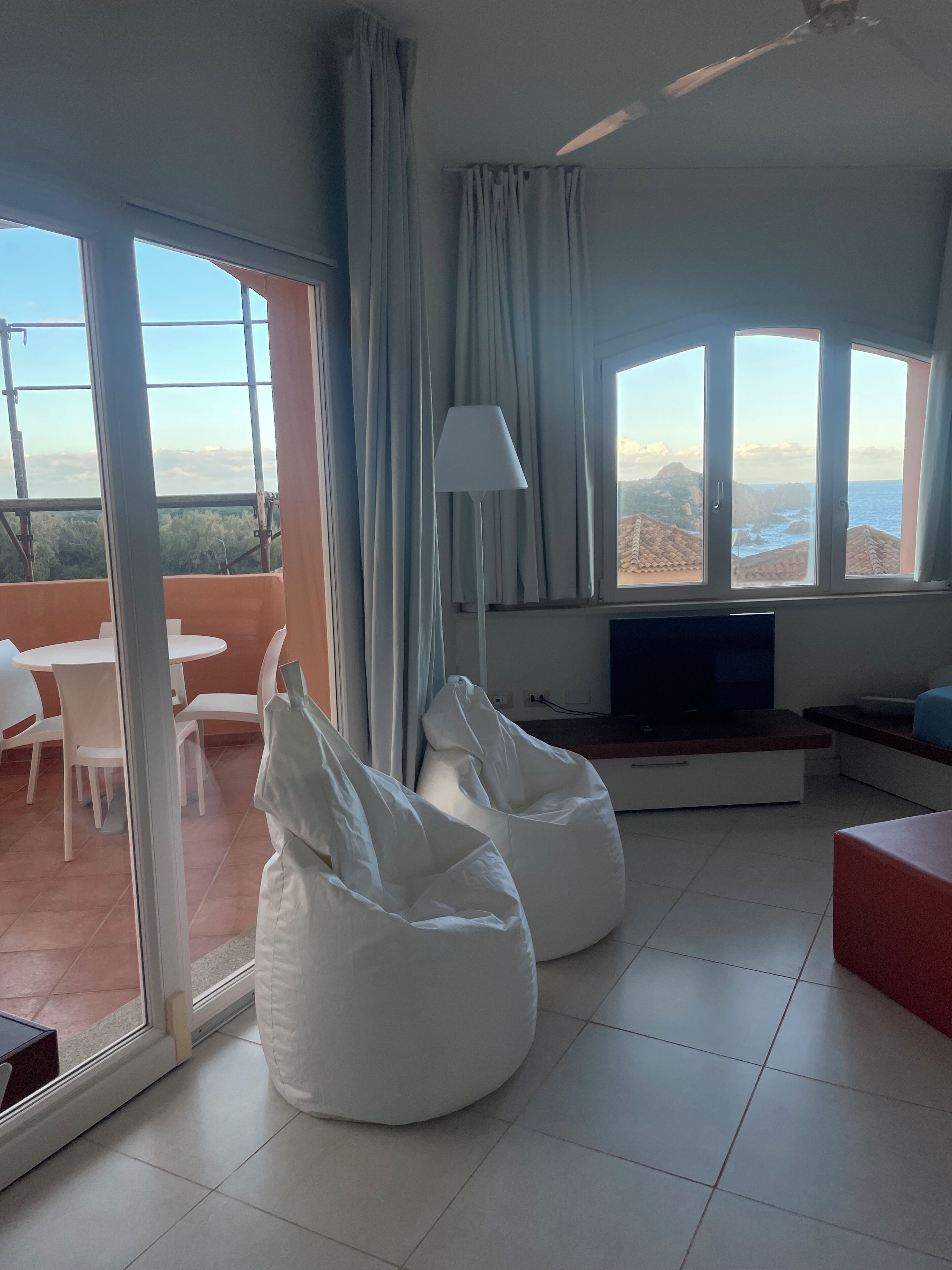Un salon moderne doté d’un téléviseur, de fauteuils en forme de poire de couleur blanche, d’un coin repas avec des chaises et de grandes fenêtres offrant une vue sur la mer.