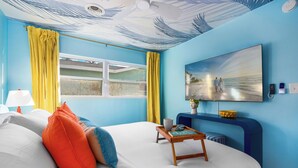 Une chambre avec un lit, une table de chevet avec une lampe, une fenêtre avec des rideaux jaunes et un tableau représentant une scène de plage accroché au mur.