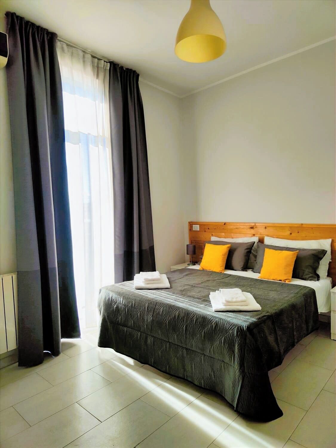Camera d'albergo con un letto grande, tende scure, una testiera in legno e una lampada a sospensione gialla.