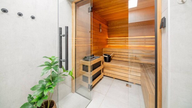 Eine moderne Sauna mit Holzwänden und einer Glastür, einer kleinen Holzbank und einem schwarzen Eimer.