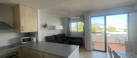 Una cocina moderna con microondas, horno y armarios de madera. Hay un comedor con mesa y sillas, y un balcón con vistas.