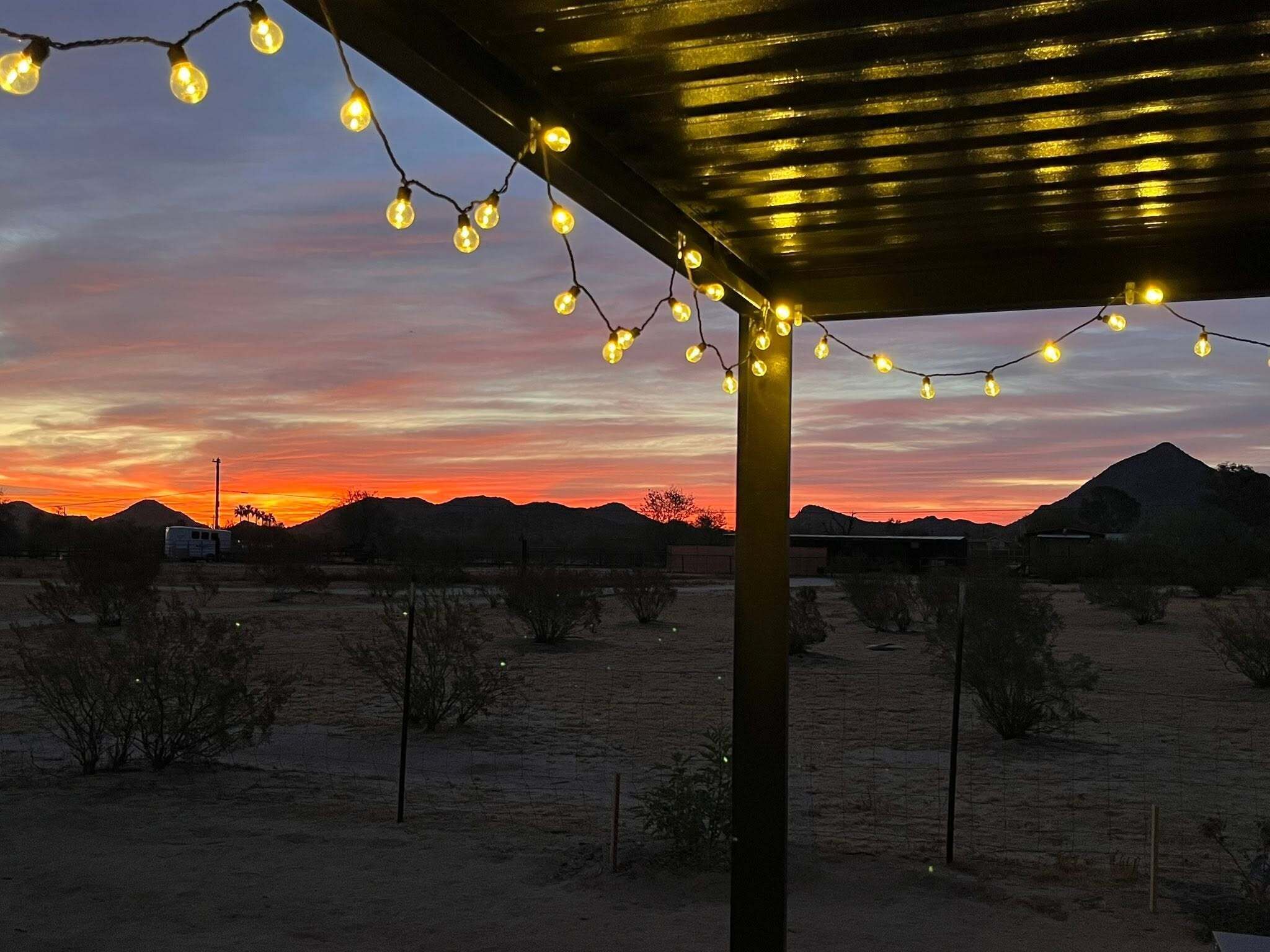 Paisaje desértico con un atardecer, una zona cubierta con luces de cuerda y montañas distantes.