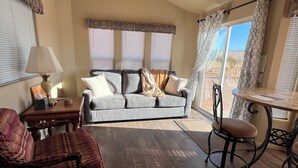 A living room with a sofa, a plaid armchair, a coffee table, and a dining table with chairs. There is a ceiling fan, a lamp, and a window with curtains.