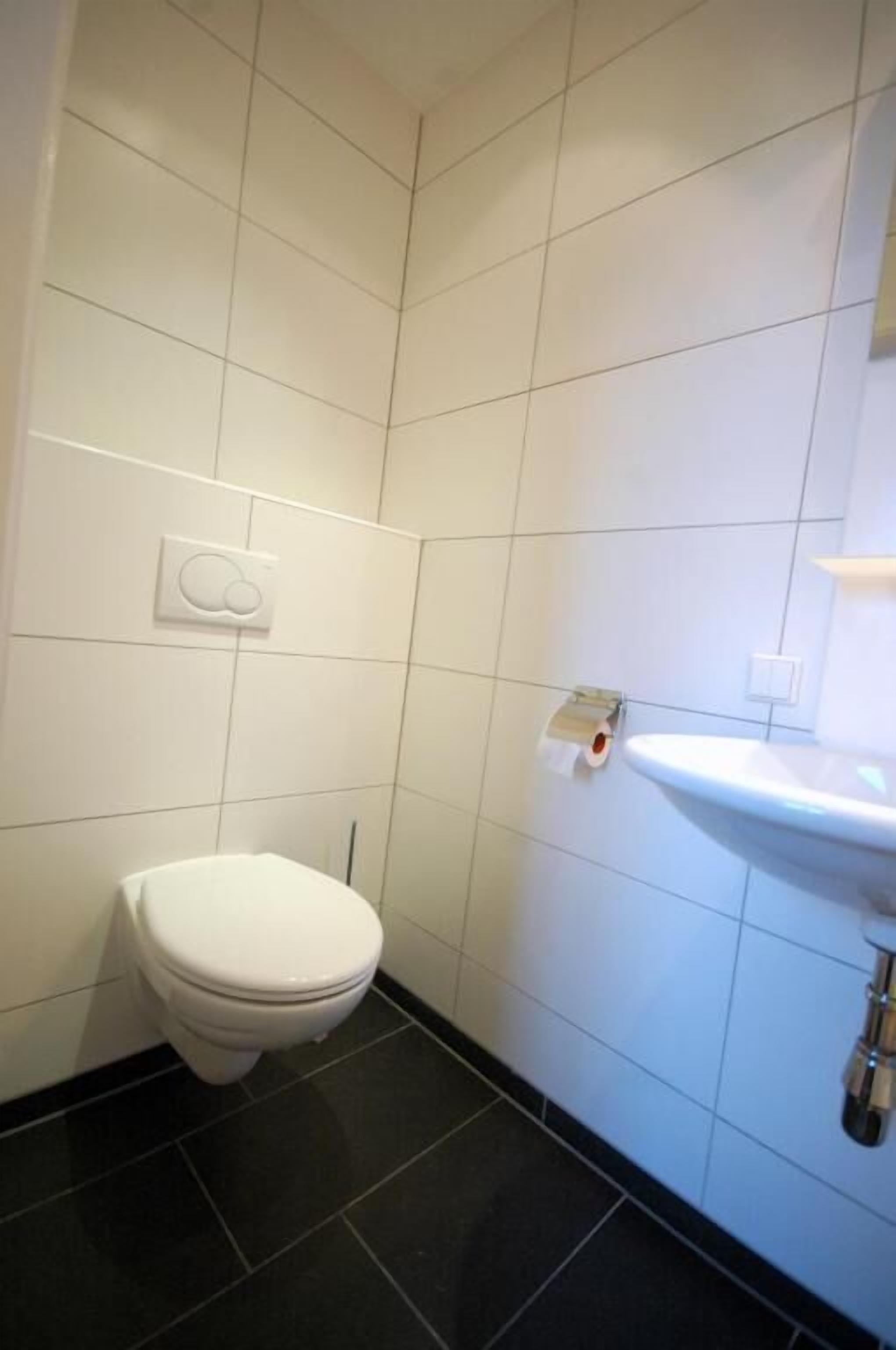 Une petite salle de bain propre, équipée de toilettes, d’un lavabo et d’un porte-rouleau de papier toilette fixé au mur.