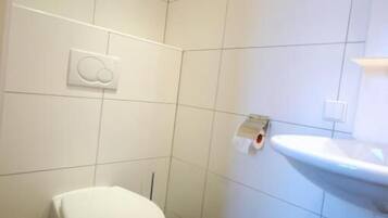 Une petite salle de bain propre, équipée de toilettes, d’un lavabo et d’un porte-rouleau de papier toilette fixé au mur.